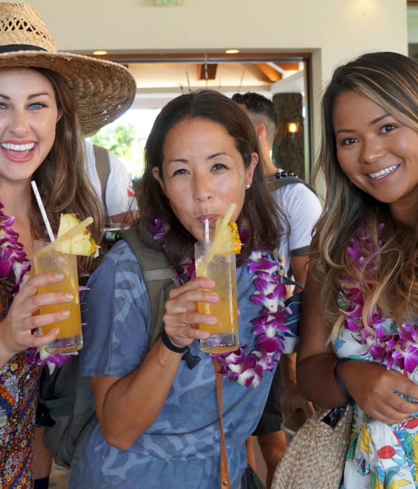 Three people wearing leis are smiling and holding tropical drinks with pineapple garnishes indoors.