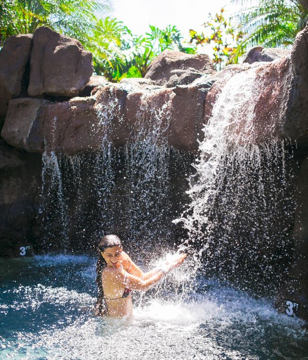 A person is enjoying a waterfall in a tropical setting, surrounded by rocks and palm trees, wearing swimwear and smiling in the water.