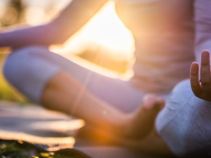 A person is meditating outdoors in the lotus position on a yoga mat, with sunlight shining through in the background, focusing on relaxation.