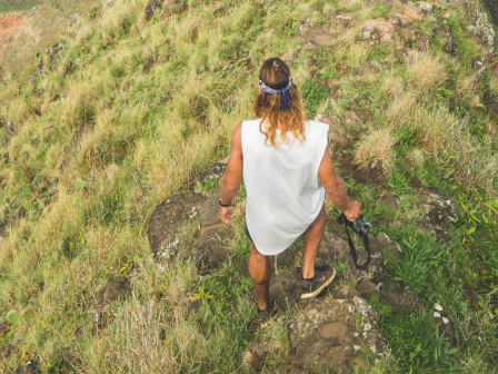 A person with long hair stands on a grassy cliff edge, wearing a casual outfit and holding a hat, overlooking a lush, green landscape below.