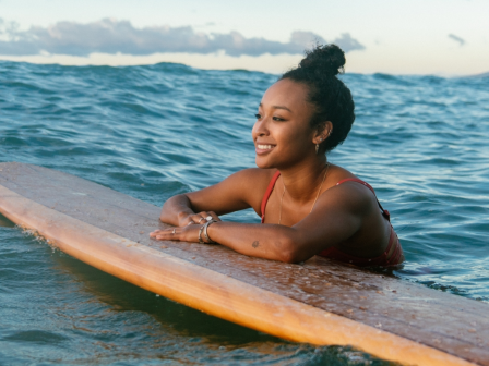 A person in a swimsuit rests on a surfboard in the ocean, with a clear sky and distant mountains in the background.