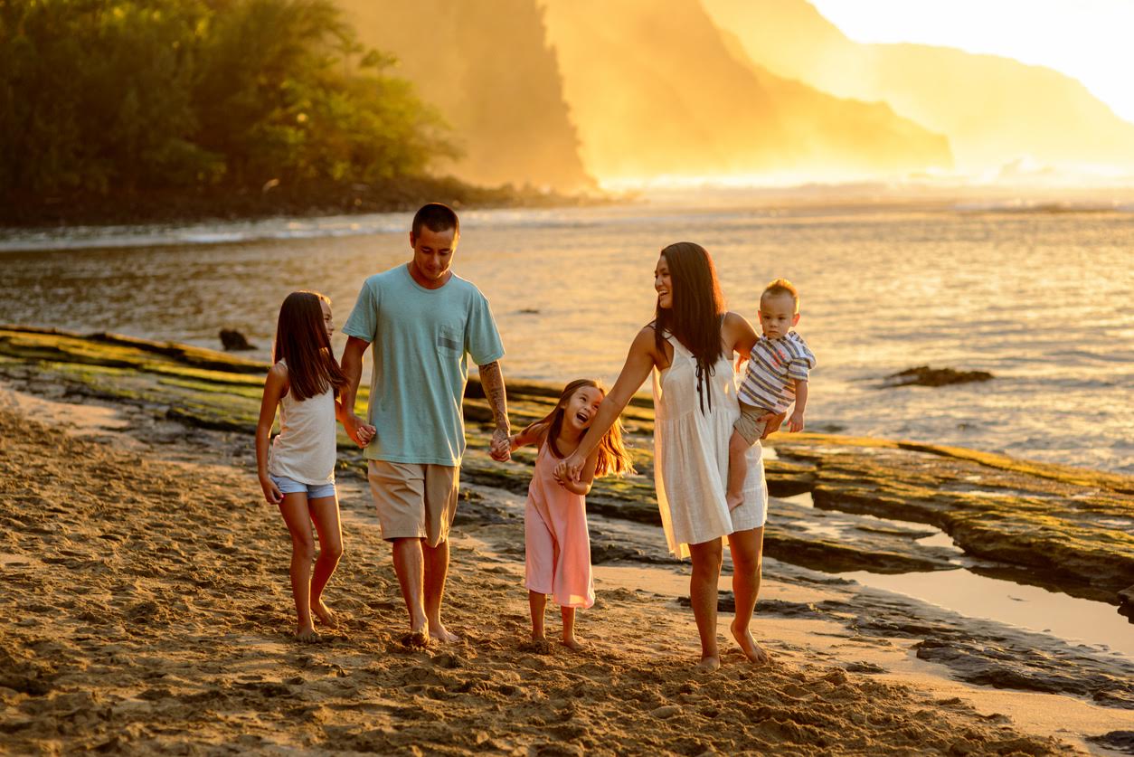A family of five walks hand in hand along a sandy beach at sunset, with cliffs in the distance and gentle waves nearby.