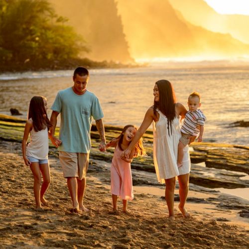 A family walks hand-in-hand along a sunlit beach at sunset, holding kids near the water.
