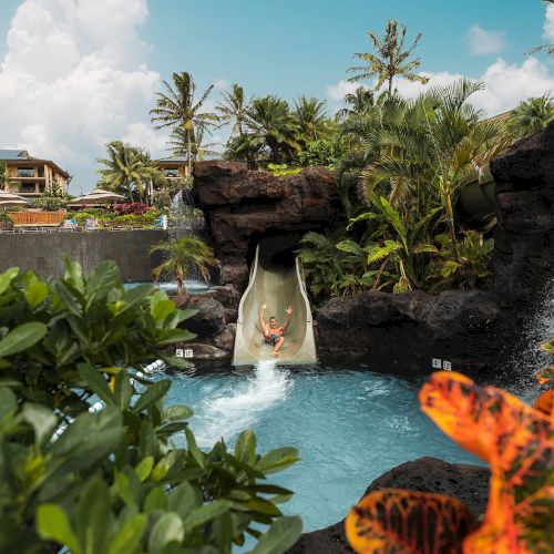 A person slides down a tropical water slide into a bright blue pool, surrounded by lush greenery and volcanic rocks, with orange flowers in the foreground.
