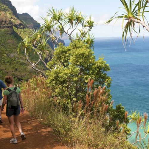 Two hikers walk along a rocky coastal trail with cliffs and blue sea to the right, sunny and clear skies.