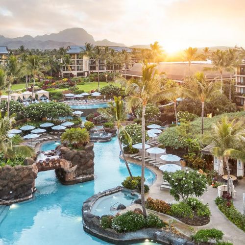 A resort-style pool with winding water slides, palm trees, lounge chairs, and sunlit buildings around a tropical oasis.