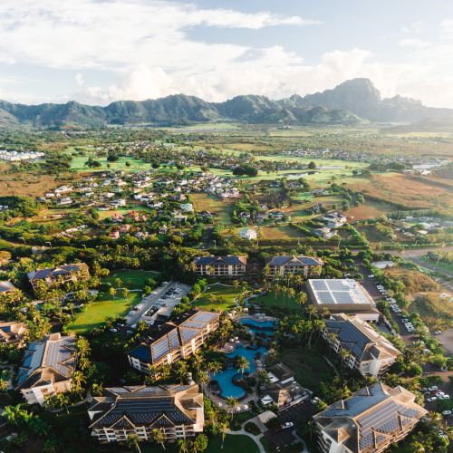 Aerial view of a resort or village with buildings, a central pool, and lush green landscape surrounded by hills and fields, under a clear sky.