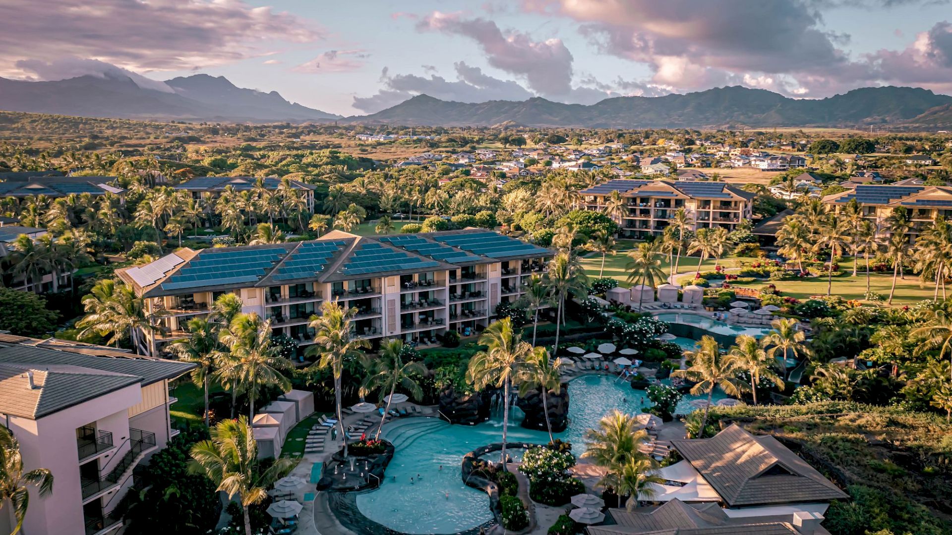 An aerial view of a resort with multiple low-rise buildings, a central curvy pool, palm trees, greenery, and distant mountains under a pink-orange sunset.