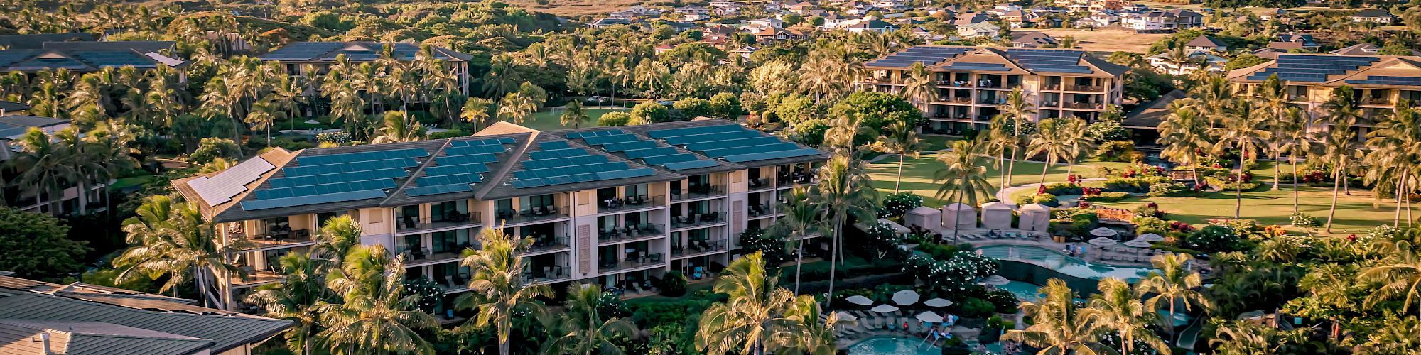 Aerial view of a tropical resort with palm trees, a curvy pool, surrounding low-rise buildings, and distant mountains under a colorful sky.