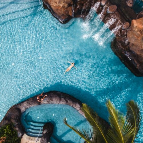 Aerial view of a resort pool with turquoise water, stepping stones, a curved seating area, palm trees, and rocky edges, invitingly sunny.