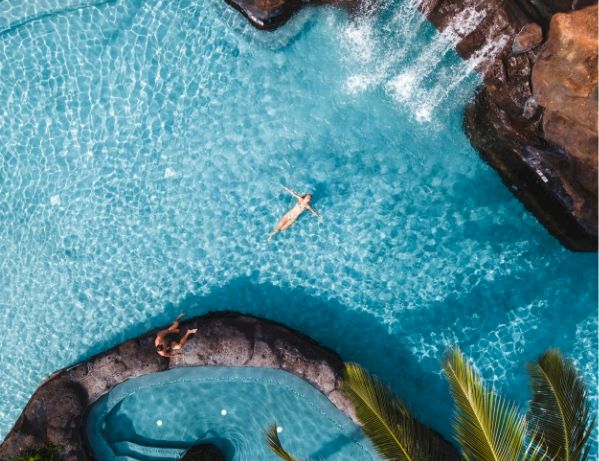 Aerial view of a resort pool with turquoise water, stepping stones, a curved seating area, palm trees, and rocky edges, invitingly sunny.