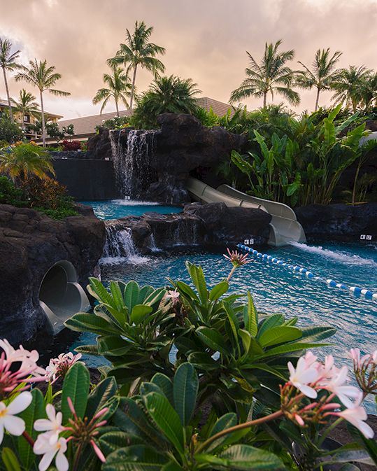 A resort pool scene with cascading waterfalls, palm trees, lush tropical plants, and a person swimming under a sunset sky.