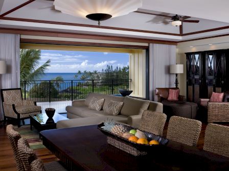 A luxurious living/dining room with a large open view of the ocean, wicker seating, a dark wood dining table, and tropical d&eacute;cor, overlooking a balcony.