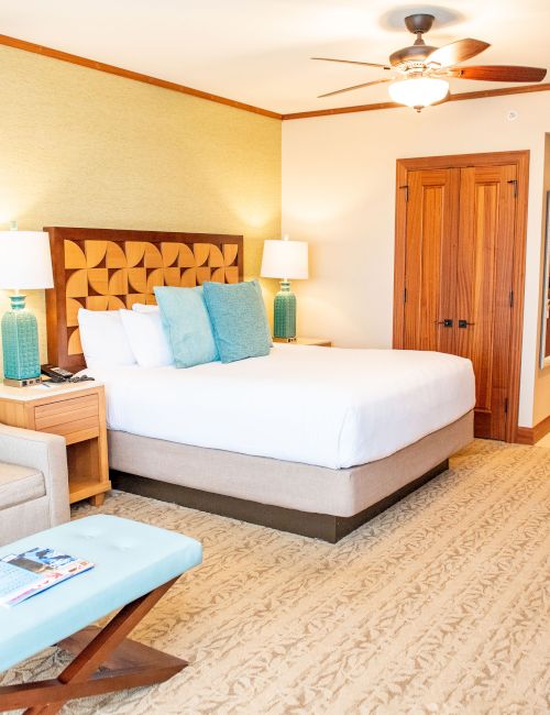 A hotel bedroom with a neatly made bed, bedside lamps, wooden doors, a ceiling fan, and a flat-screen TV.