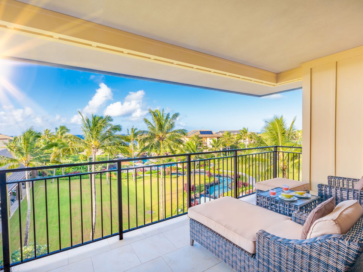A sunny apartment balcony with wicker seating and a view of palm trees, a pool, and bright blue sky with clouds.
