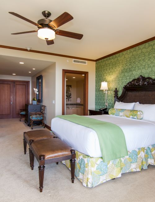 A hotel-style bedroom with a canopy headboard, green patterned walls, bedside lamps, vanity area, and a ceiling fan above.