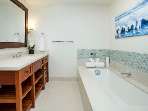 A bright bathroom with a wooden vanity, mirrors, rolled towels, a bathtub, and blue wave wall art above tiled walls.