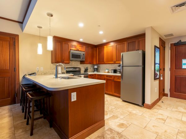 A modern kitchen with wood cabinets, a stove, microwave, granite island, sink, and a stainless refrigerator.