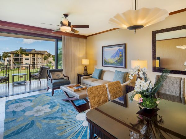Bright living room with an ocean-view balcony, sofa and coffee table, ceiling fans with lamps, framed art, and orchids.