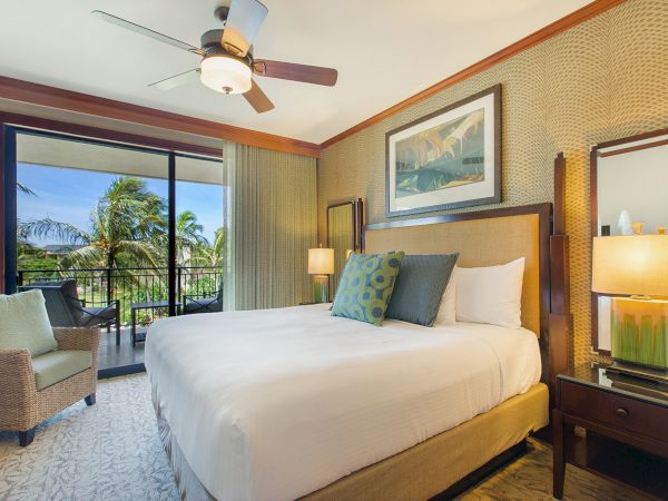 A sunny hotel bedroom with a king bed, patterned walls, bedside lamps, a ceiling fan, and a balcony view with tropical palms.