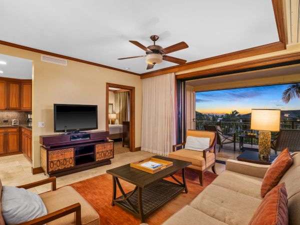 A cozy living room with a ceiling fan, TV, sofa and coffee table, plus open balcony view and a kitchenette with wooden cabinets.