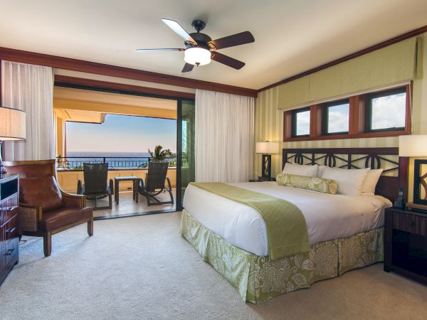 A hotel bedroom with a king bed, ceiling fan, striped curtains, and lounge chairs facing the ocean view.