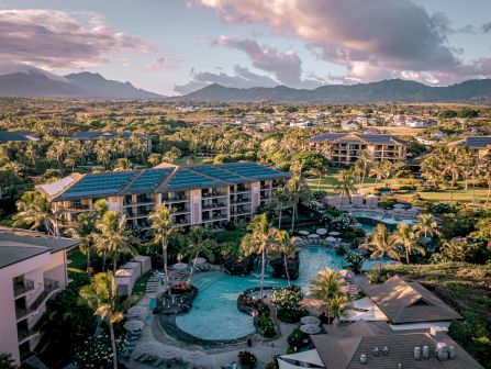 A tropical resort with multi-story buildings, rooftop solar panels, a palm-lined pool, and mountains at sunset.