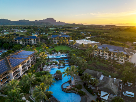 An aerial view of a tropical resort at sunset with palm gardens, a large blue swimming pool, and solar panels beside mountains.