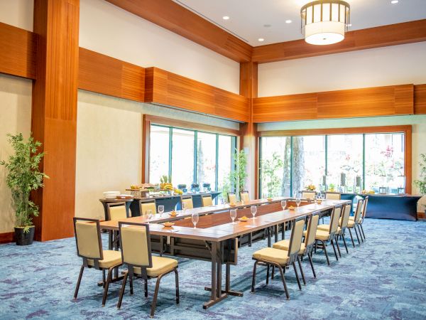 A modern conference room with a long dining table, cushioned chairs, pendant lights, large windows, bamboo plants, and buffet setup.