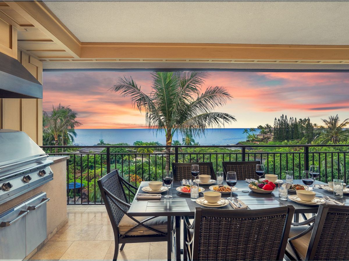 A seaside balcony dining set with wine, a grill, and palm trees overlooking the ocean at sunset.