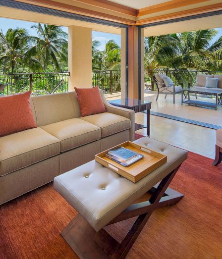 A bright tropical balcony living room with beige sofas, coffee table, and chairs, looking out over lush palm trees.
