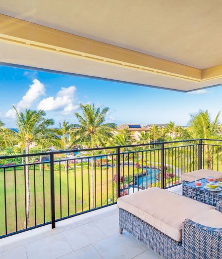 Sunlit balcony with wicker sofa and table, overlooking palm trees, a pool, and blue sky over sunny houses.