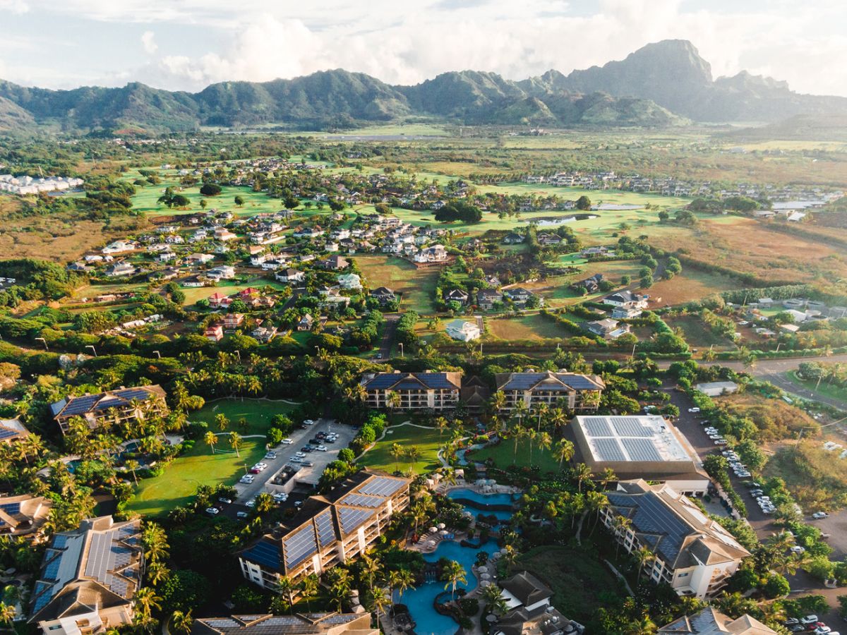 Aerial view of a tropical resort and village with pools, solar roofs, fields, and mountains under a bright cloudy sky.