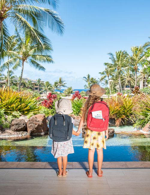 Two travelers in sun hats with red and black backpacks stand by a tropical pool, surrounded by palms and lush greenery.