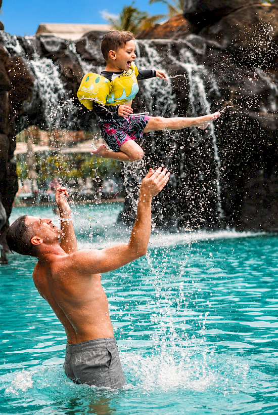 A man tosses a child from a pool water slide, and they splash into the sunny turquoise water with droplets flying.