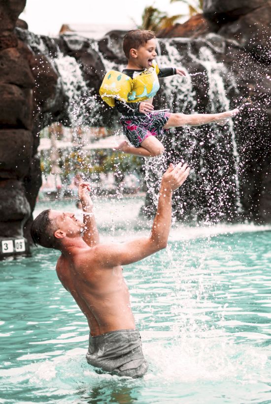 A man throws a child into a pool, creating a huge splash in a sunny rock-and-waterplay area.