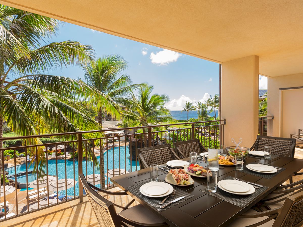 Tropical resort balcony with a set outdoor dining table, palm trees, a blue pool, and ocean view under sunny skies.