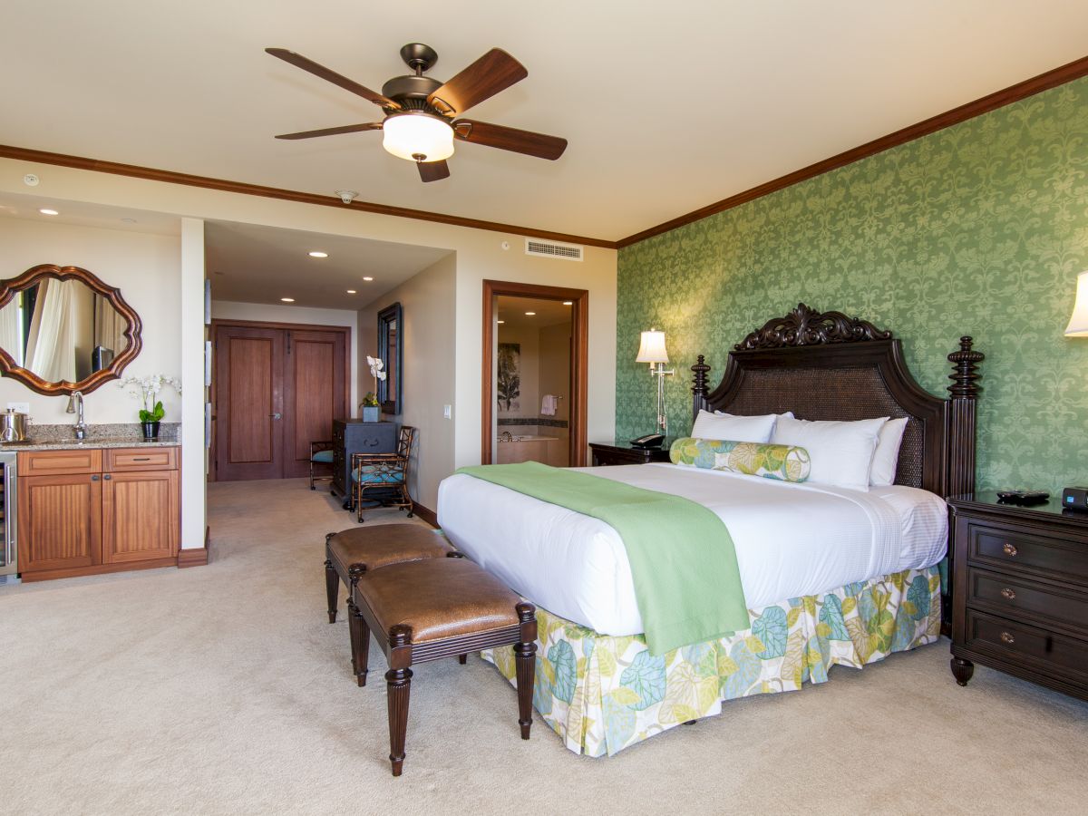 A hotel bedroom with a large bed, green patterned wallpaper, ceiling fan light, bedside lamps, and a vanity area with mirror.