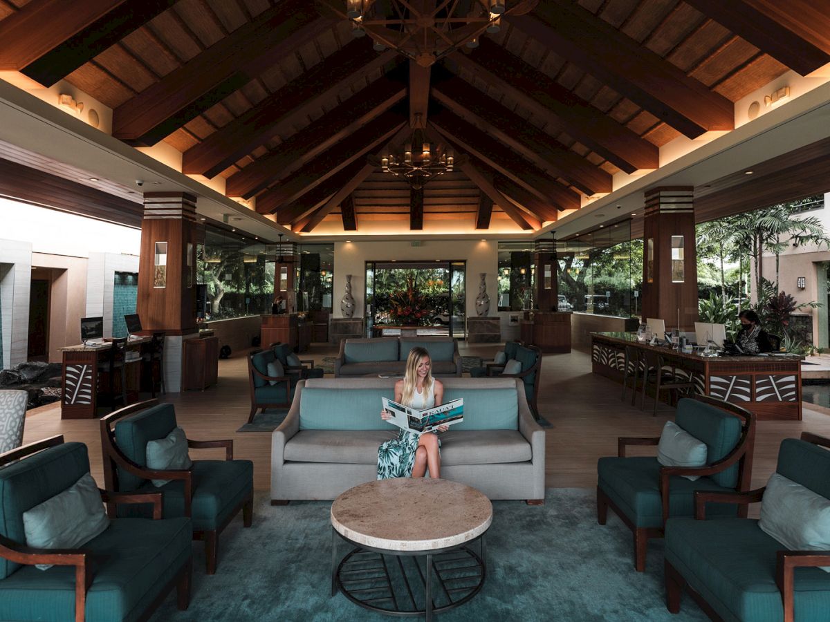 A luxurious lounge with a vaulted wooden ceiling, chandeliers, teal sofas, and a woman relaxing and reading a magazine.