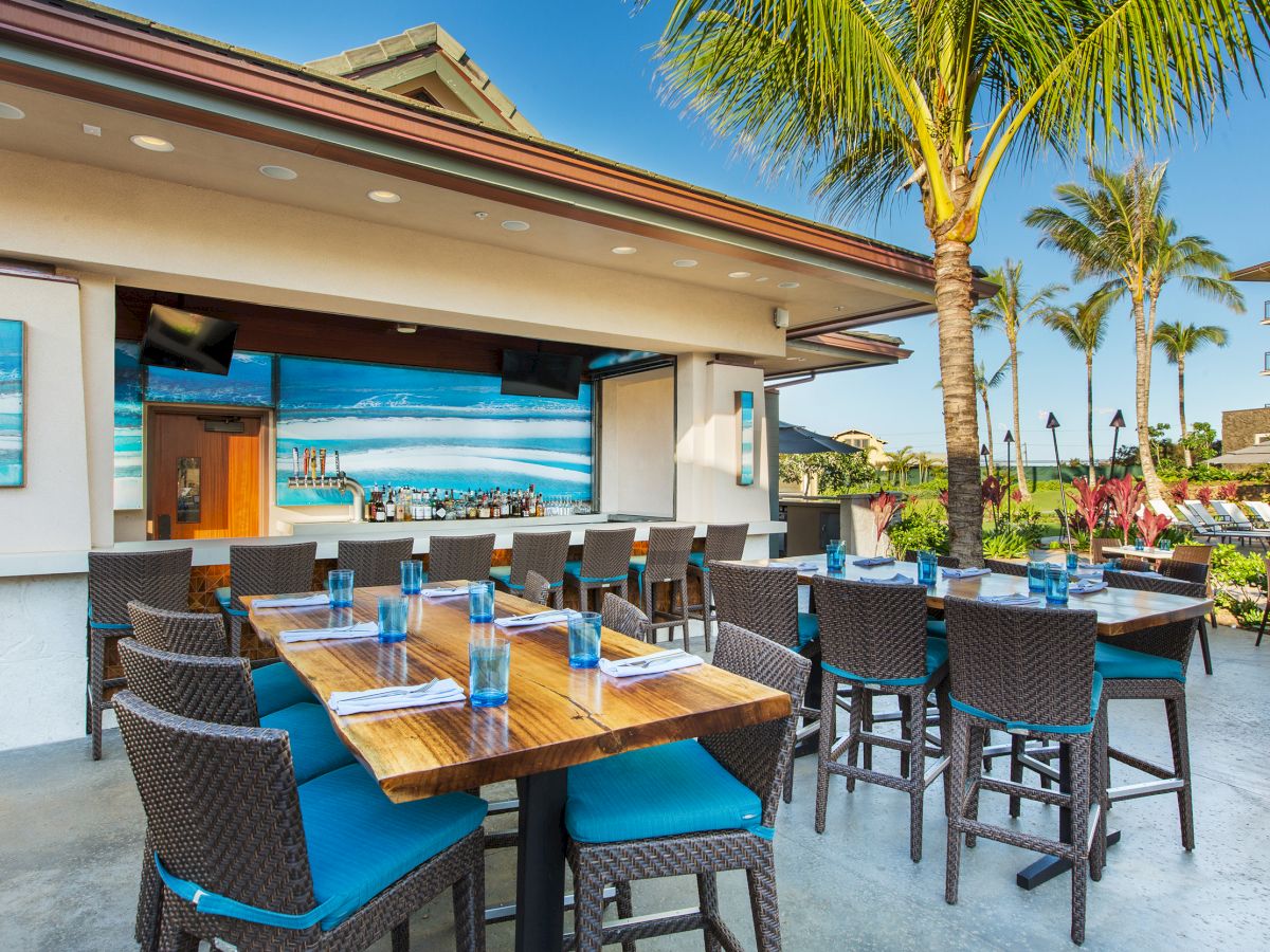 Tropical hotel patio with a covered bar, wooden tables, lounge seating, and palm trees under a bright blue sky.