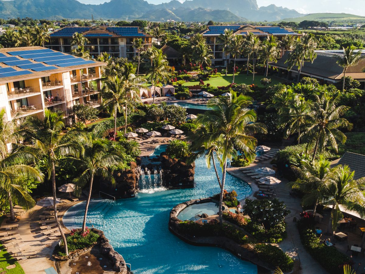A tropical resort with palm trees, a winding pool, solar panels, and distant mountains under a bright blue sky.