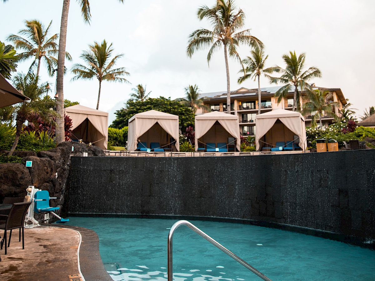 Tropical resort scene with a pool, palm trees, and tan cabanas against a cloudy sky.