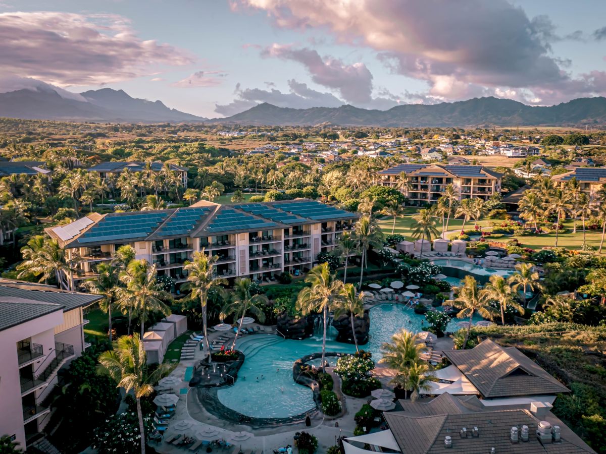 A tropical resort with a large outdoor pool, palm trees, solar panels on buildings, and a sunset view of mountains in the distance.
