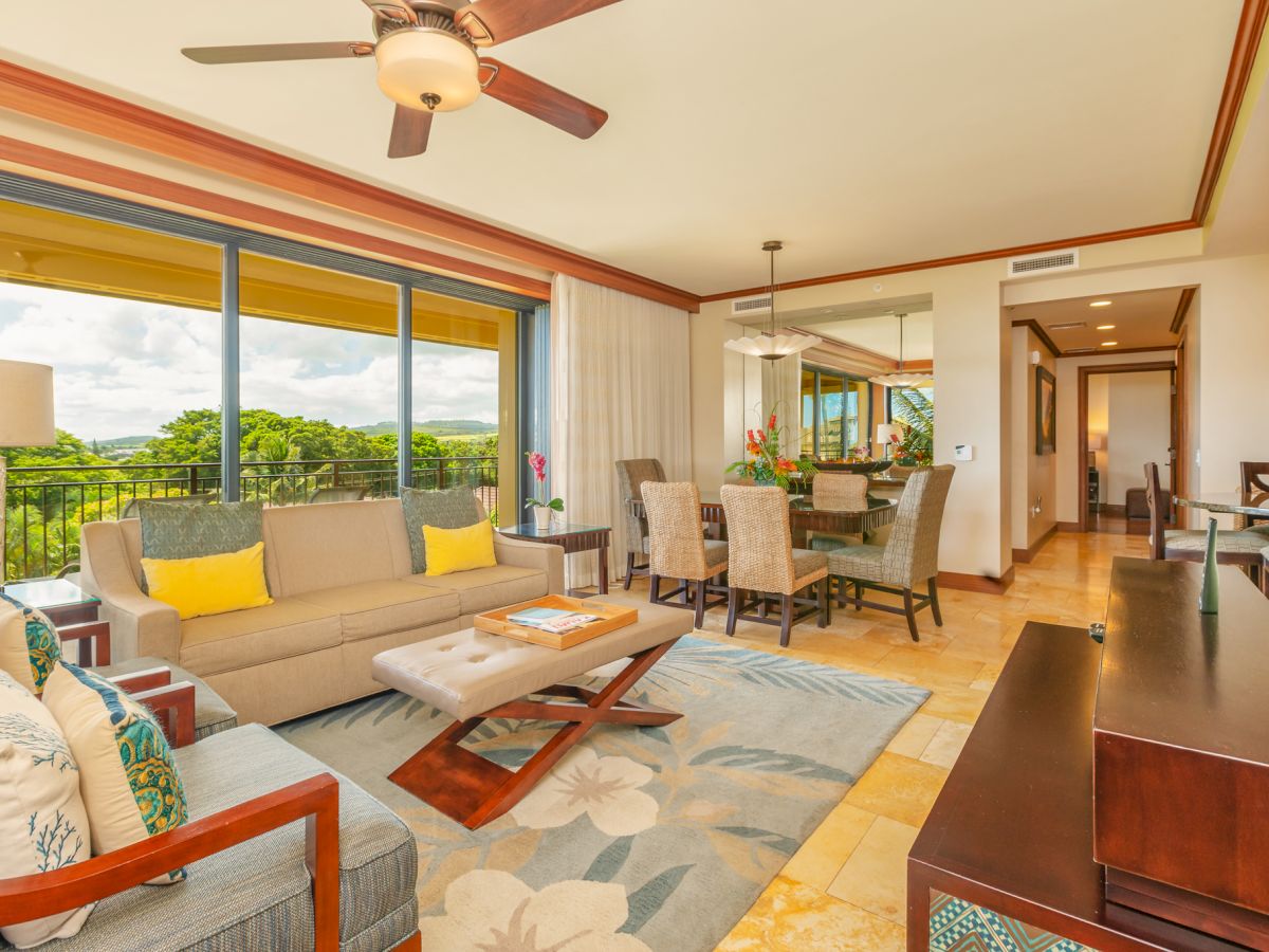 A bright tropical living/dining room with a sofa, dining chairs, large windows, and a ceiling fan.