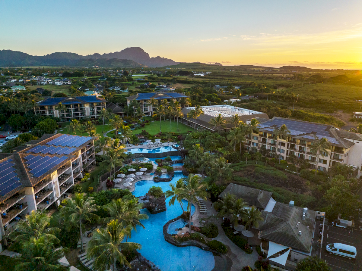 Aerial view of a tropical hotel resort with swimming pools, palm gardens, solar panels, and mountains at sunset.