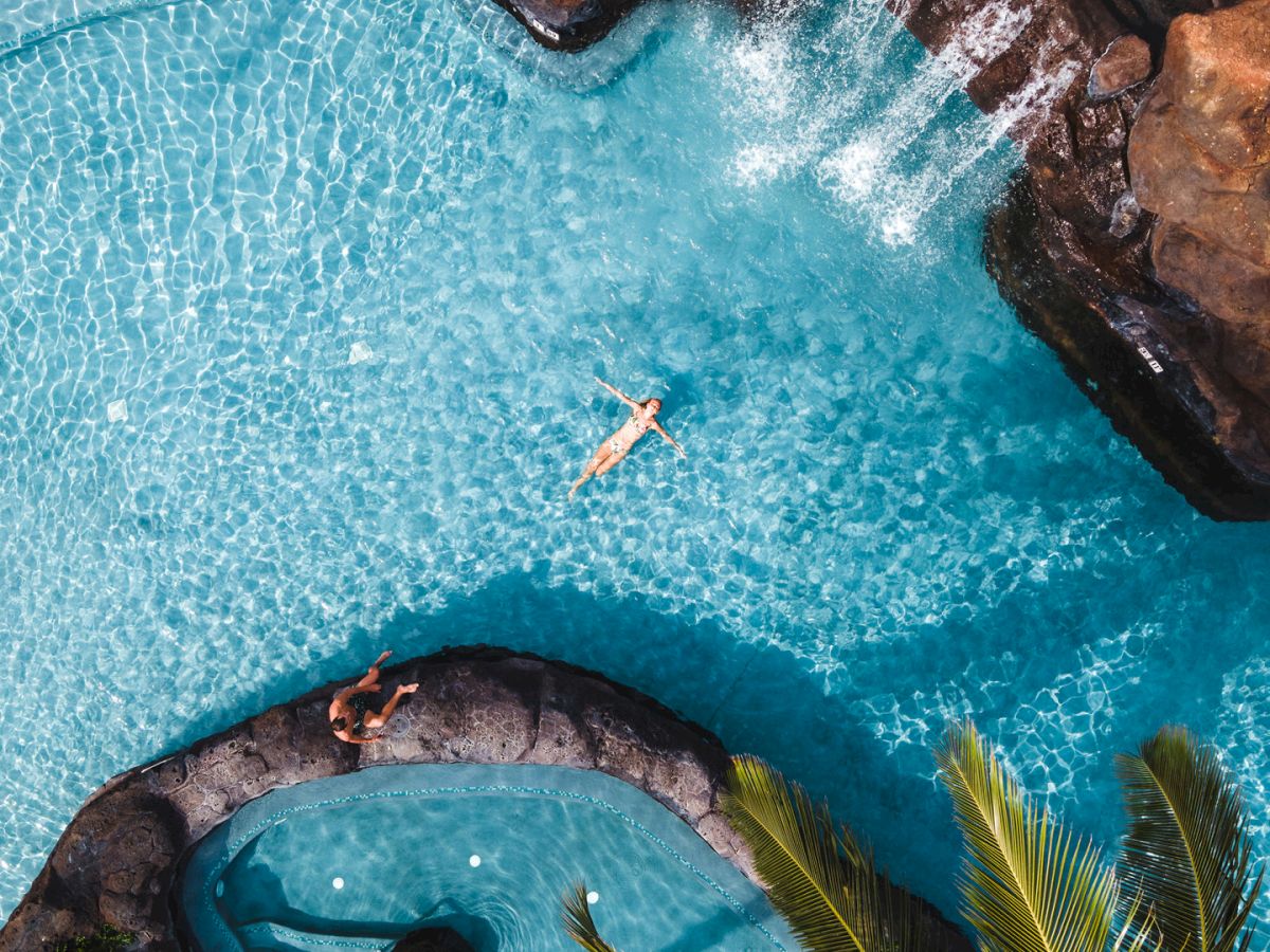 Aerial view of a bright infinity pool beside rocky cliffs, with a swimmer in clear blue water and palm leaves framing it.