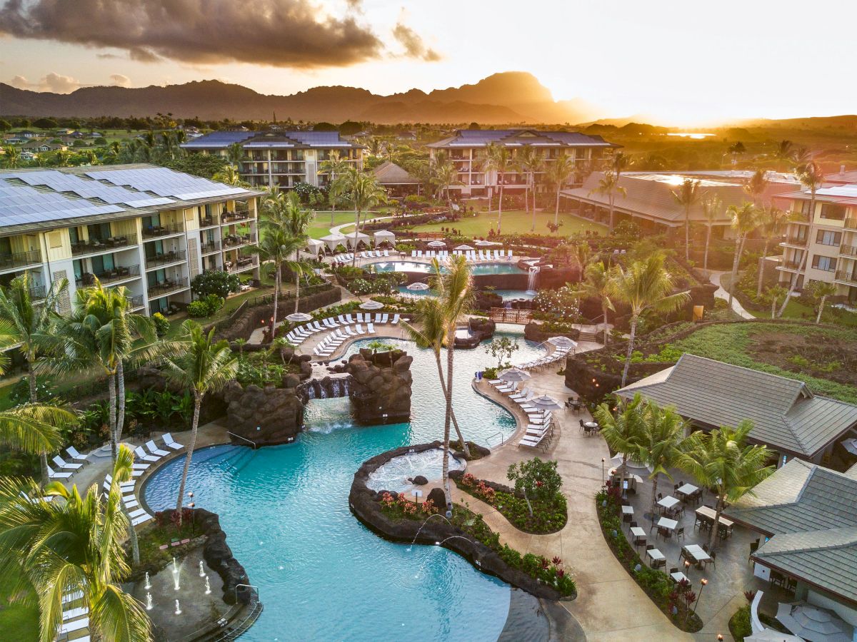 A tropical resort at sunrise with palm trees, a curved pool, sunlit buildings, and mountains in the background.