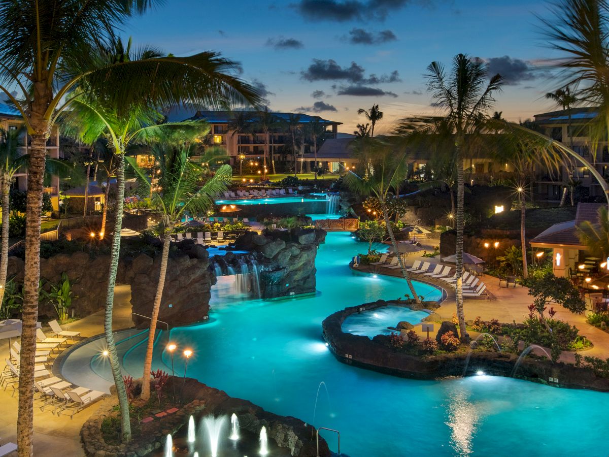 A tropical resort at dusk with palm trees, glowing buildings, and a large illuminated pool with waterfalls.