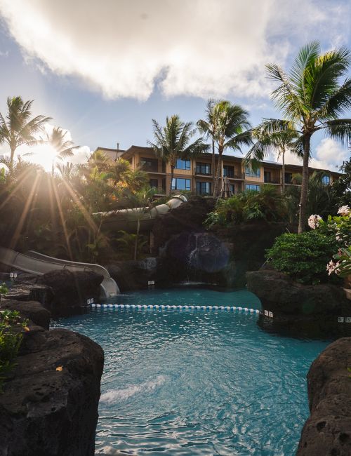 Tropical resort pool with turquoise water, palm trees, sun rays, and a hotel building under a partly cloudy sky.