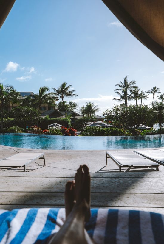A resort poolside view with blue water, sun loungers, umbrellas, and palm trees under a bright sky.